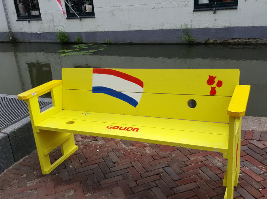 Knallgelbe Sitzbank, bemalt mit niederländischer Flagge und der Beschriftung "Gouda", vor schmalem Wasserkanal. Anschließend noch ca. 2 Meter der angrenzenden Häuserfront sichtbar.