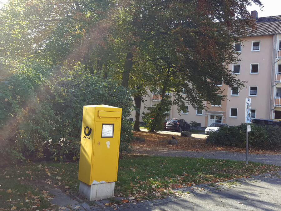 Im Vordergrund ein großer gelber Postkasten, der auf einem Rasenstück steht, das zu einem parkähnlichen Gelände vor einer Mehrfamilien- Häuserzeile gehört.